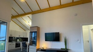 Ein modernes Wohnzimmer mit einem Wand-Fernseher, einer Holzkabine mit Glastüren und einem Essbereich mit Tisch und Stühlen.