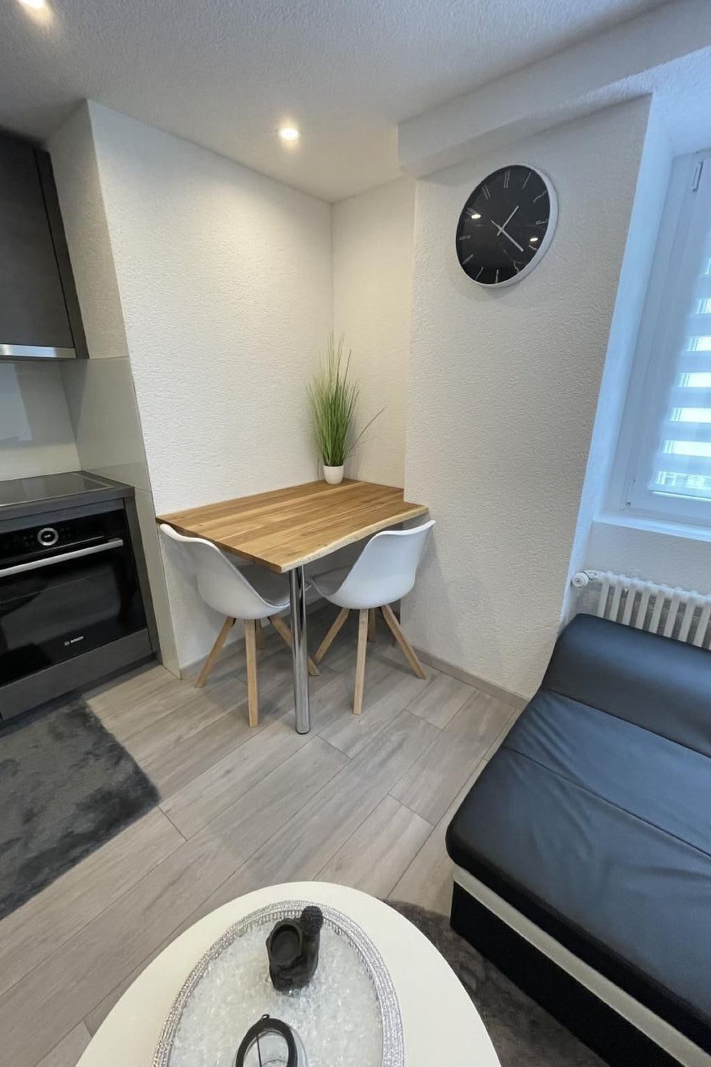Un espace de vie compact comprenant un coin cuisine, une table à manger avec deux chaises, un canapé et une horloge murale.