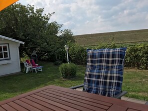 Eine Terrasse mit einem Holztisch, einem karierten Kissen, einem weißen Stuhl und einem kleinen, weißen Gebäude mit einem Fenster.