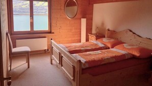 Une chambre confortable avec un plafond en bois, une grande fenêtre donnant sur un lac, un miroir rond, une table de chevet et un lit avec une literie à motifs floraux.