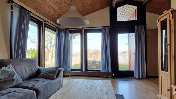 Ein geräumiges Wohnzimmer mit Holzboden, einem großen Fenster mit Vorhängen, einem grauen Sofa und einem Holztisch.