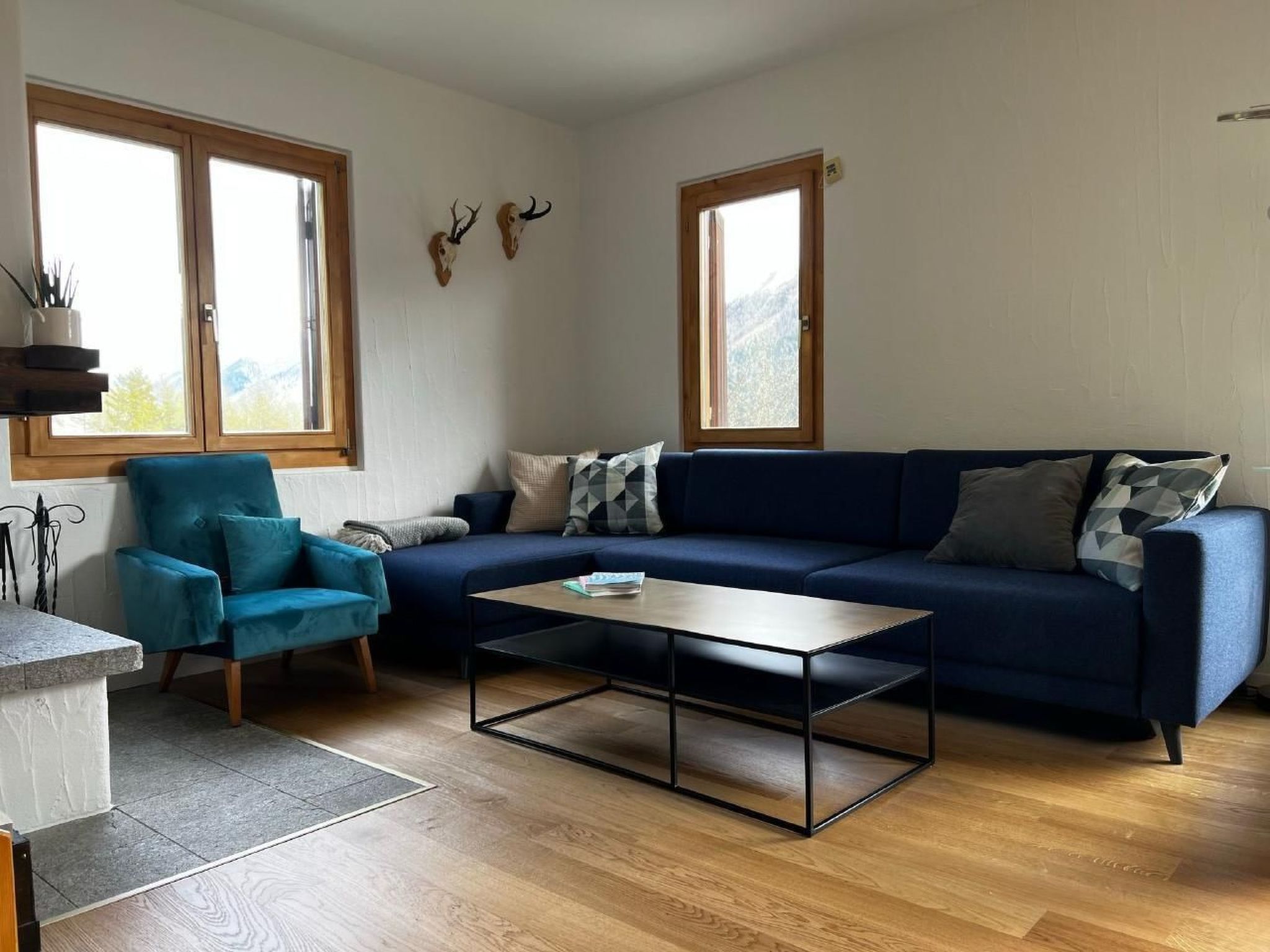 Ein Wohnzimmer mit einem blauen Sofa, einem türkisgrünen Sessel, einem Couchtisch und zwei Fenstern mit Holzrahmen.