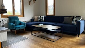 Una sala de estar con un sofá azul, un sillón verde azulado, una mesita de centro y dos ventanas con marcos de madera.