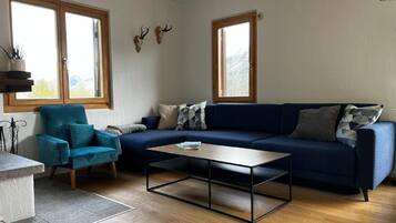 Ein Wohnzimmer mit einem blauen Sofa, einem türkisgrünen Sessel, einem Couchtisch und zwei Fenstern mit Holzrahmen.