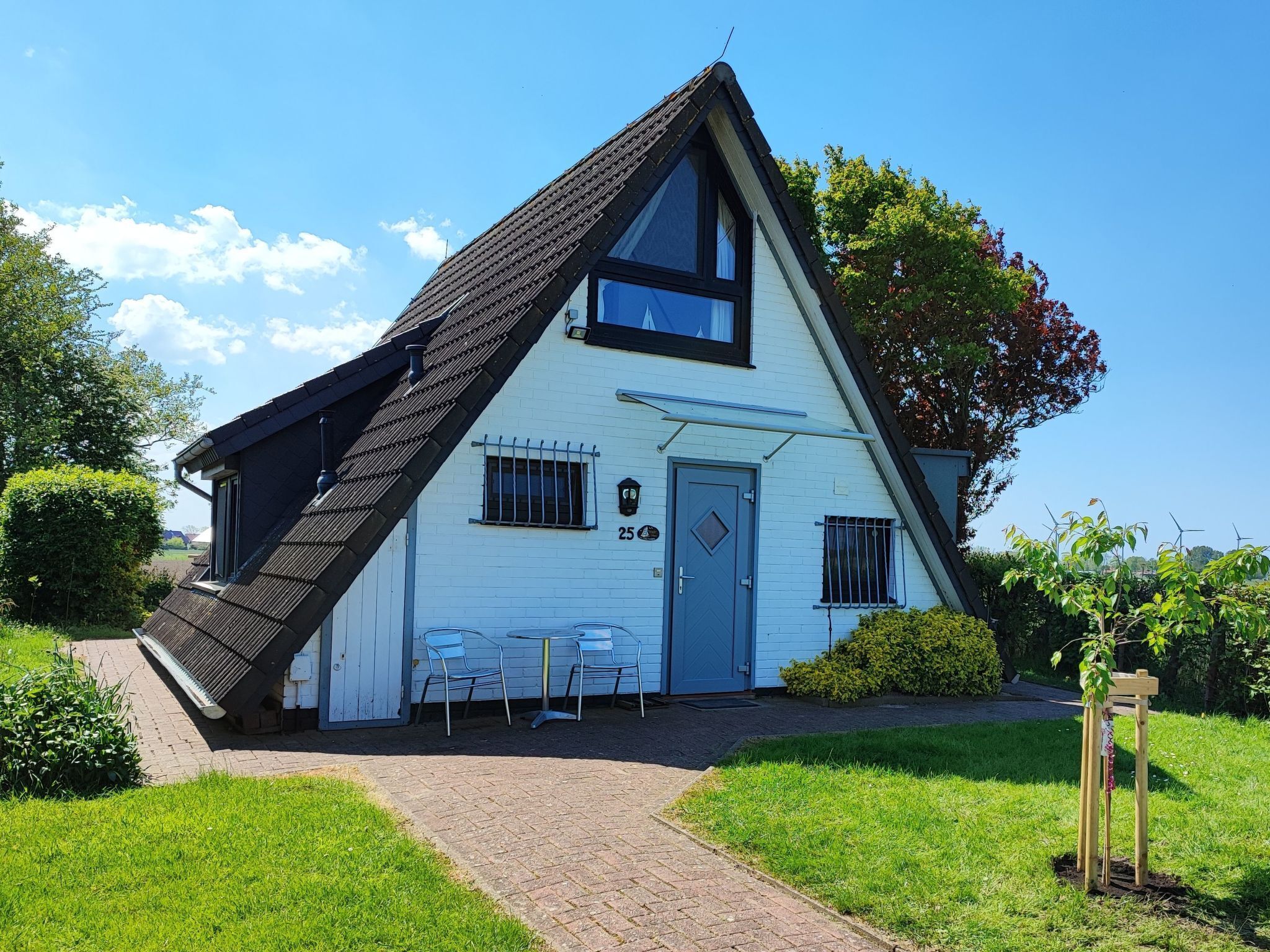A white house with a steeply pitched roof, a small porch with chairs, and a number '25' on the door.