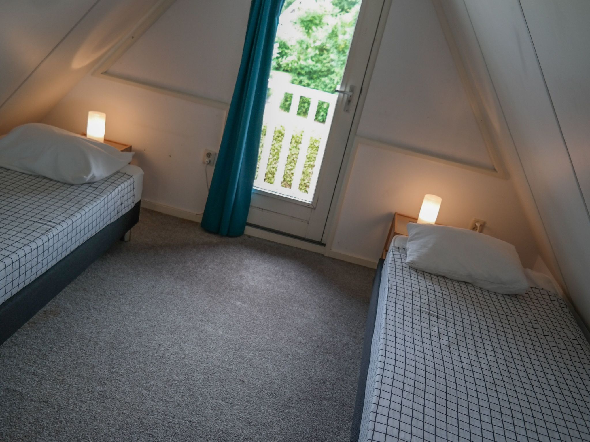 Pequeña habitación triangular con dos camas individuales, cada una con un almohadón blanco y una lámpara sobre una mesita de noche. Una cortina color turquesa cuelga cerca de una ventana con vistas a la vegetación.