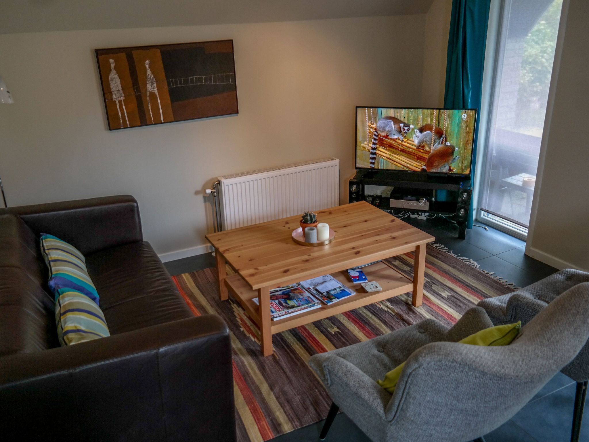 Ett vardagsrum med en brun lädersoffa, ett träbord med tidningar, en tv på ett stativ och ett fönster med gardiner.
