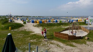 Una playa con arena, agua y gente. Hay tiendas de campaña y un parque infantil.