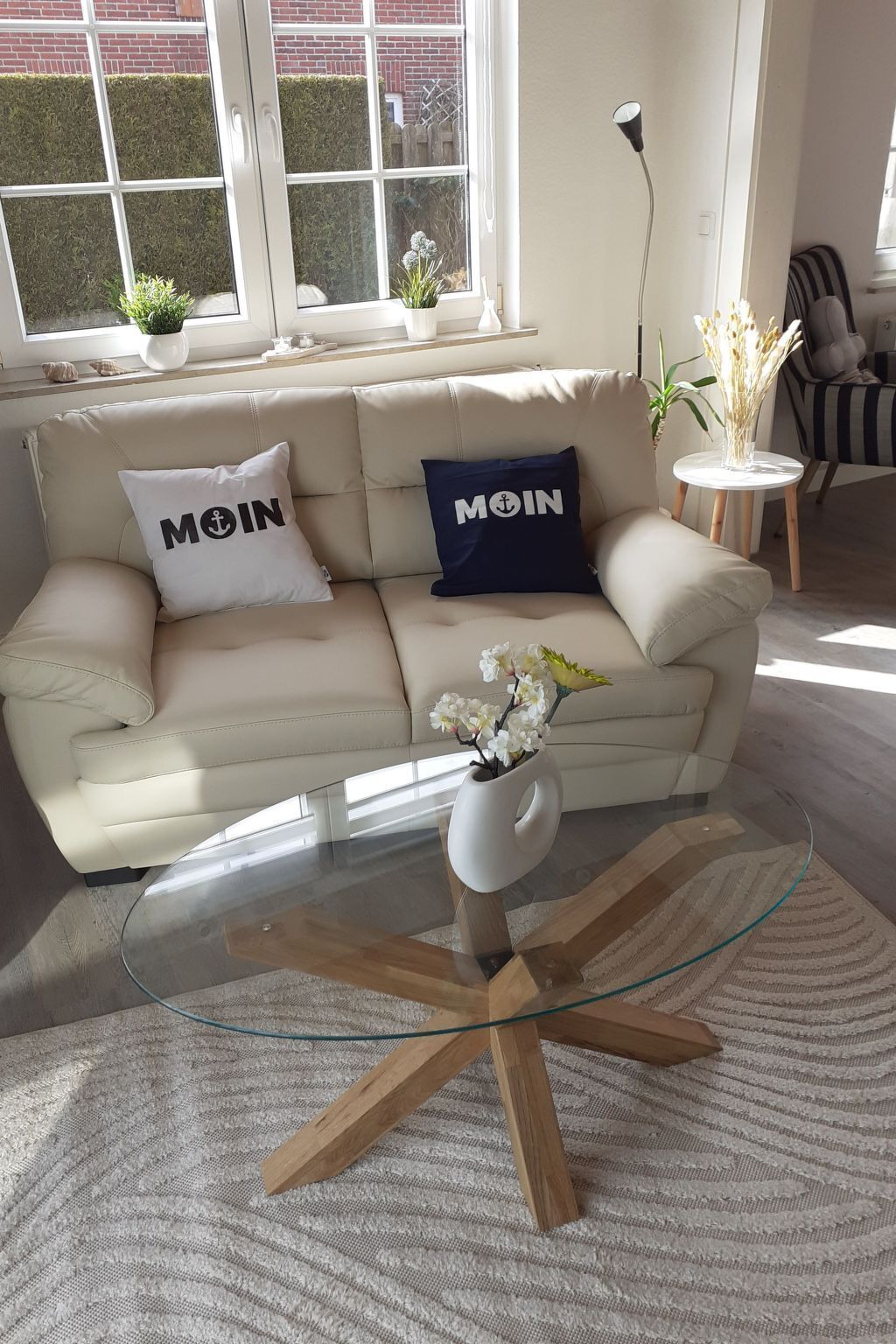 A living room with a beige sofa, two pillows with "MOIN" text, a glass coffee table, and a vase with flowers.