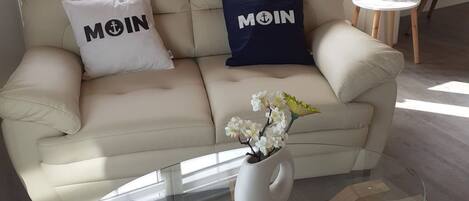 A living room with a beige sofa, two pillows with "MOIN" text, a glass coffee table, and a vase with flowers.