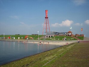 A beach with a large water tower, a building with a red roof, and a grassy area with people. - Nettes Ferienhaus in Großwürden mit Garten by Interhome (Burhave)