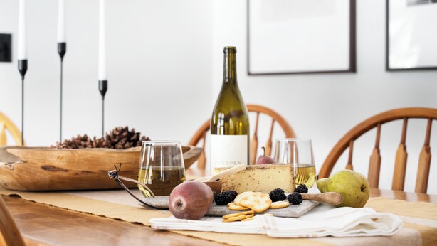 Ett träbord med en vinflaska, glas, ost, frukt och nötter.