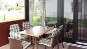Een terras buiten met een houten tafel en geruite stoelen, grote ramen en uitzicht op de omgeving.