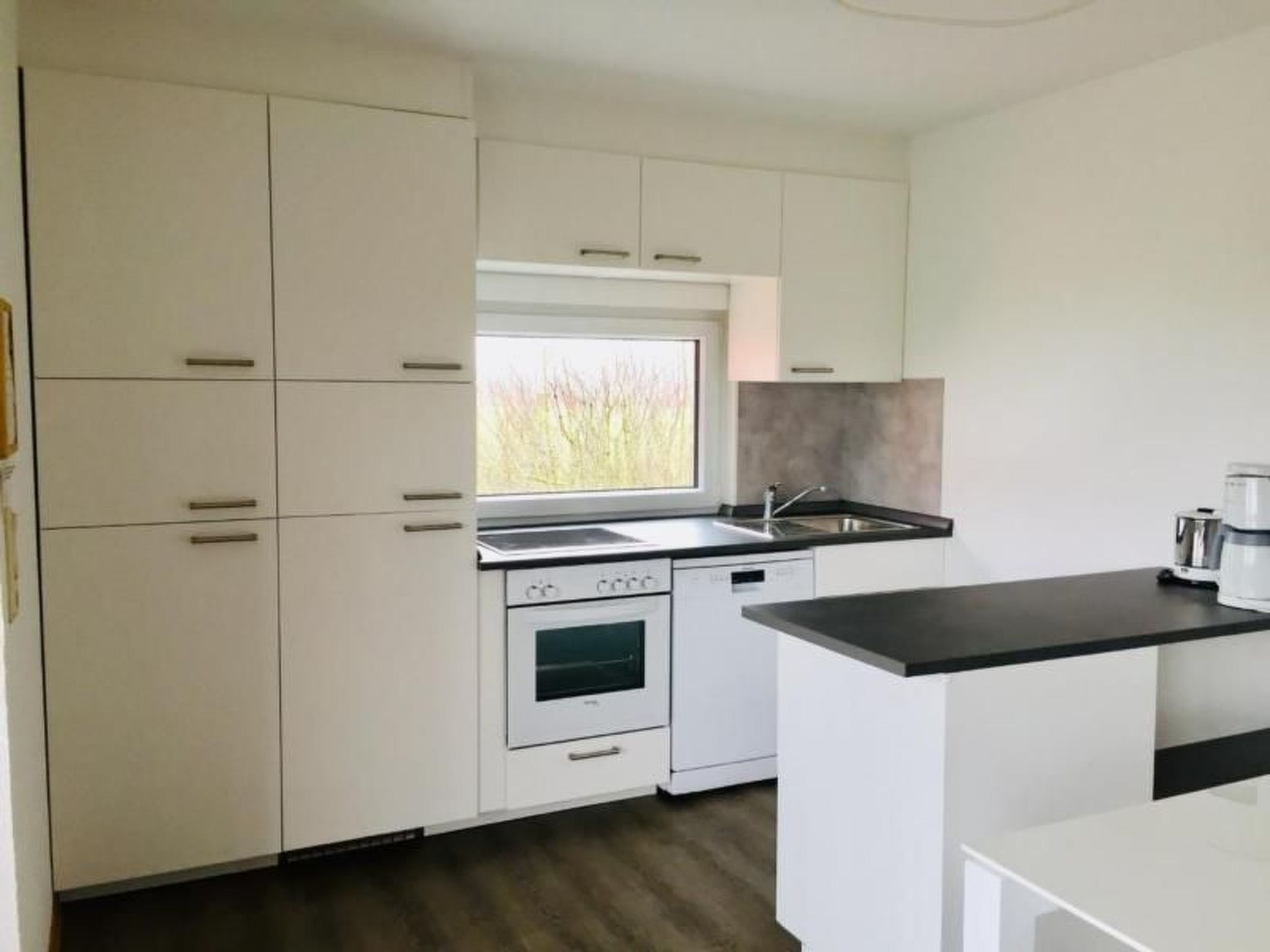 Eine moderne Küche mit weißen Schränken, einem eingebauten Backofen, einer Spülmaschine und einem Fenster, das für natürliches Licht sorgt.
