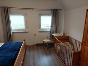 Ein Schlafzimmer mit einem Bett, einem Kinderbett, einem Heizkörper und zwei mit Vorhängen versehenen Fenstern.