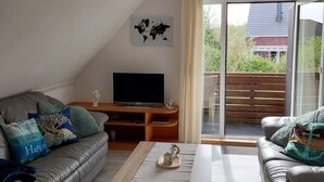 Un salon moderne avec un canapé, une table basse et un téléviseur. Une carte du monde est accrochée au mur et une porte coulissante offre une vue sur l’extérieur.