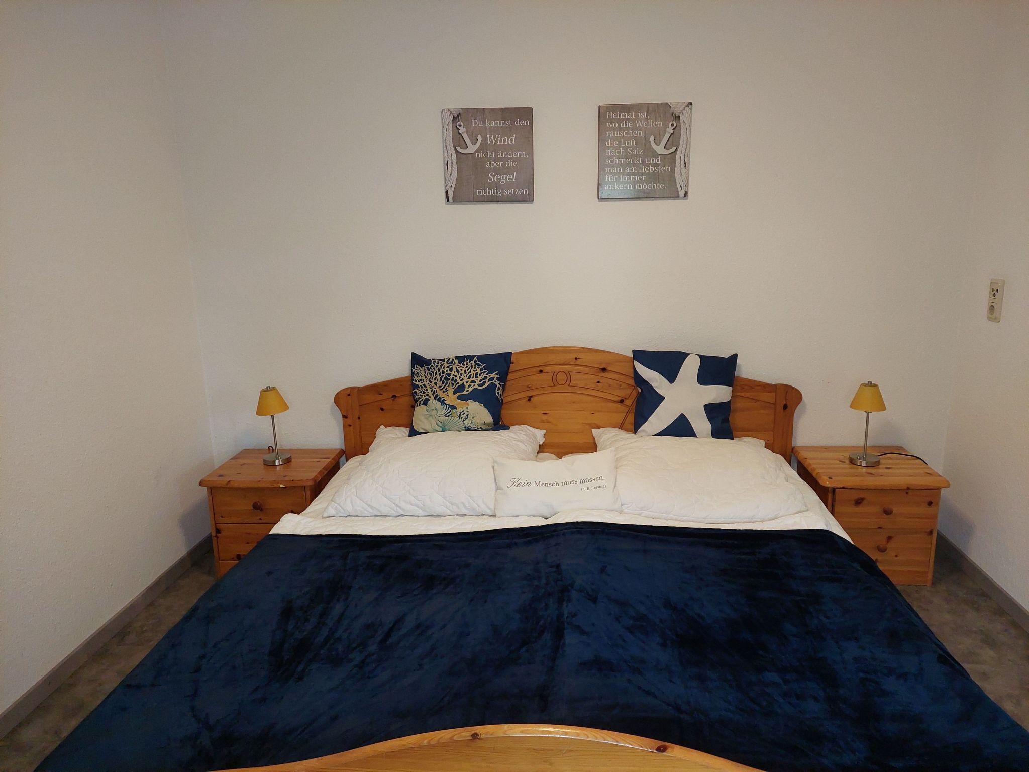 A bedroom with a wooden headboard, two bedside tables with lamps, a bed with white and blue bedding, and two wall-mounted signs with German text.