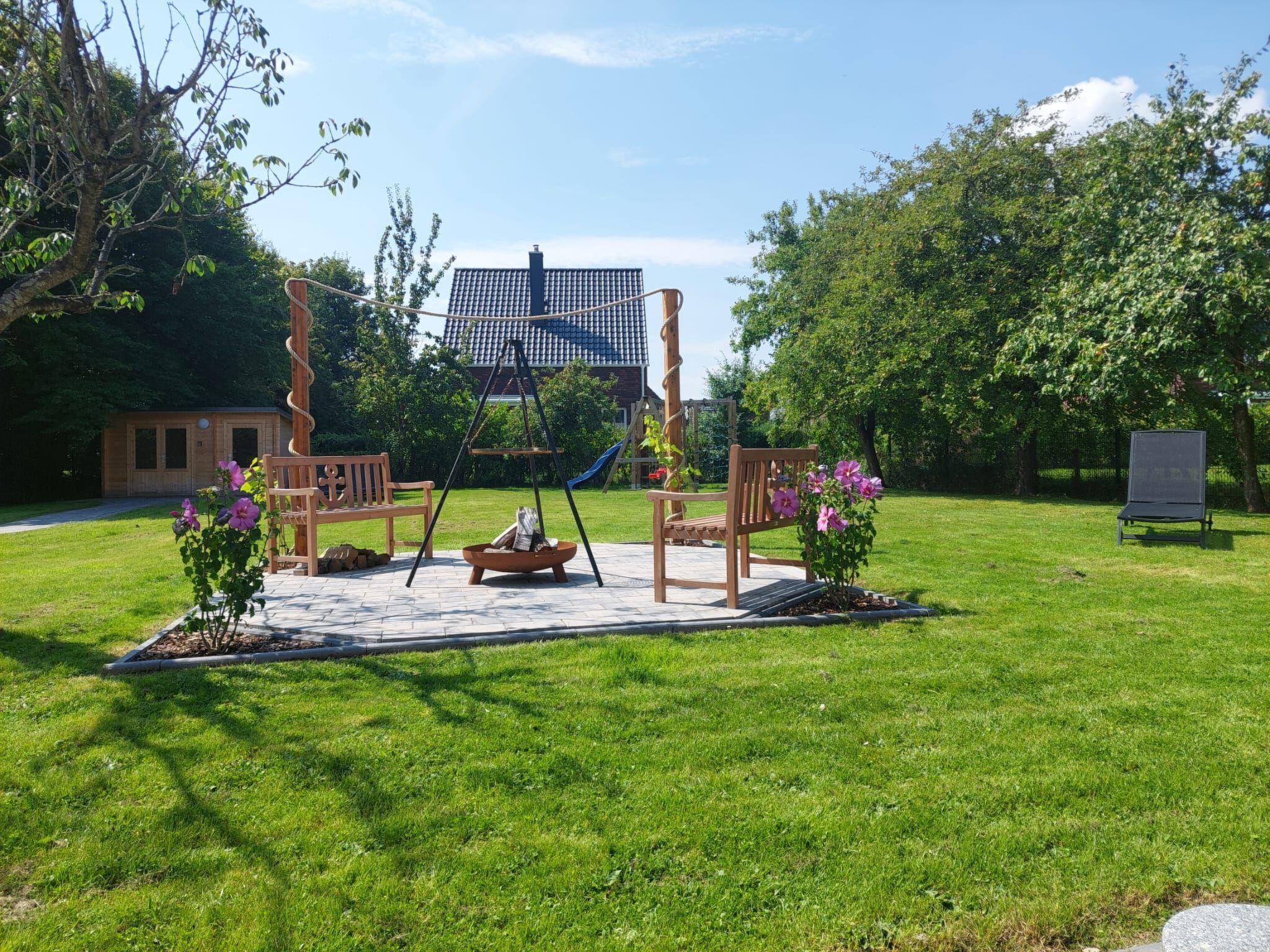 A wooden swing set with a bench and a small table in a grassy area with trees and a building in the background.
