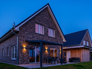 Een tweelaags huis met een bakstenen gevel, een overdekte patio met tuinmeubilair en een grote schuifdeur die leidt naar de binnenruimte.
