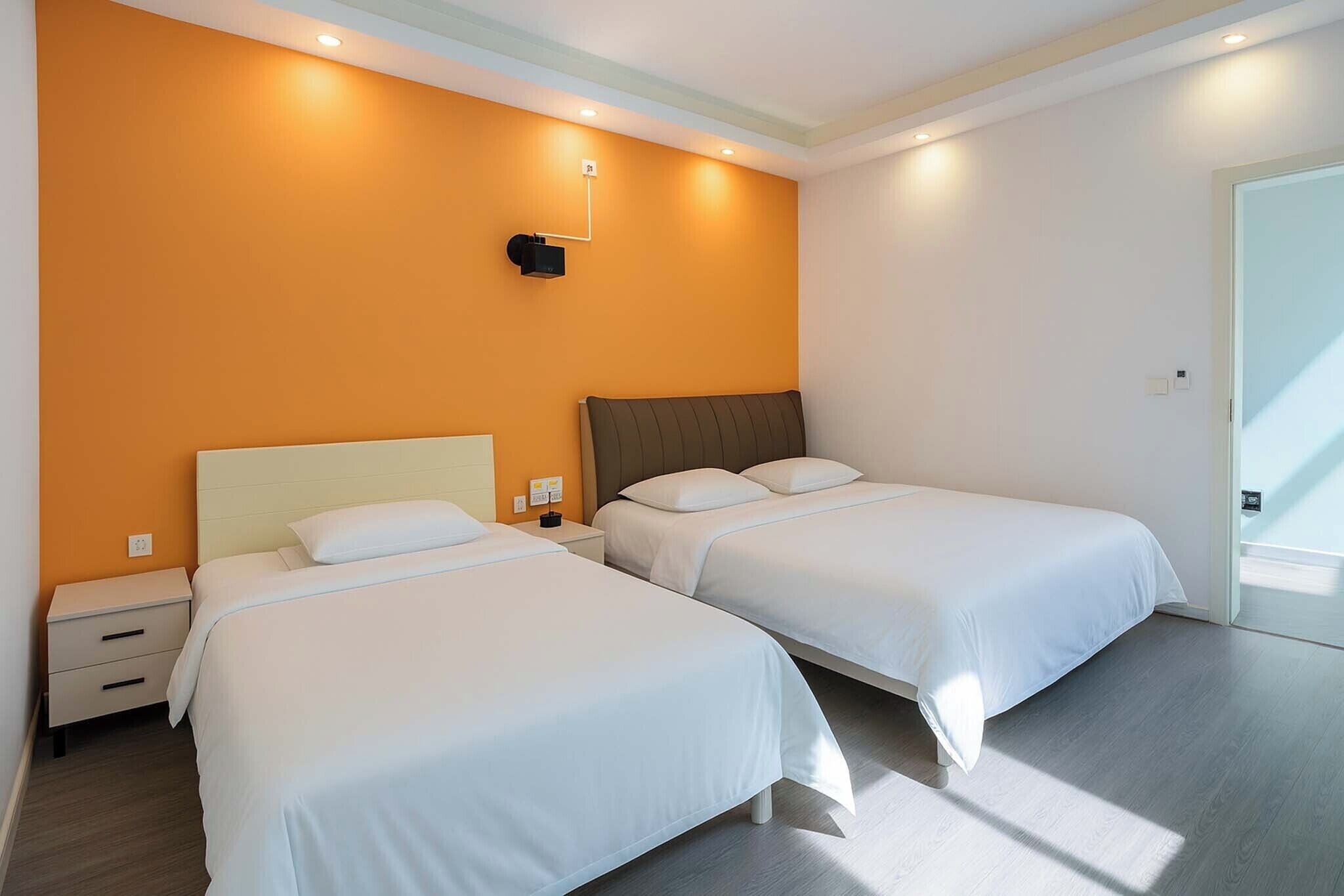 Quarto de hotel com duas camas de solteiro, uma mesinha de cabeceira, uma luminária fixada na parede e uma porta à direita.