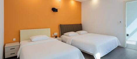 Quarto de hotel com duas camas de solteiro, uma mesinha de cabeceira, uma luminária fixada na parede e uma porta à direita.