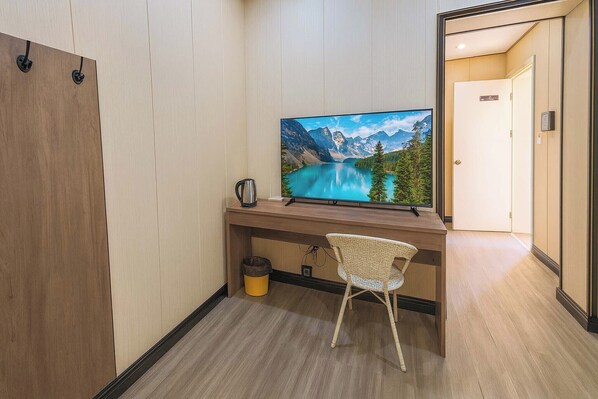 A hotel room with a wooden desk, a television displaying a mountain lake, a wicker chair, and a door leading to another room. - Tianya No.7 Homestay (Sanya)