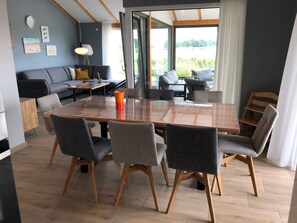 Ein moderner Essbereich mit einem großen Holztisch, grauen Polsterstühlen und Blick ins Freie durch große Fenster.