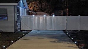 Une allée fermée par un portail mène à une maison dotée d’un garage, qui est elle-même entourée d’une clôture et éclairée par des lampes extérieures.