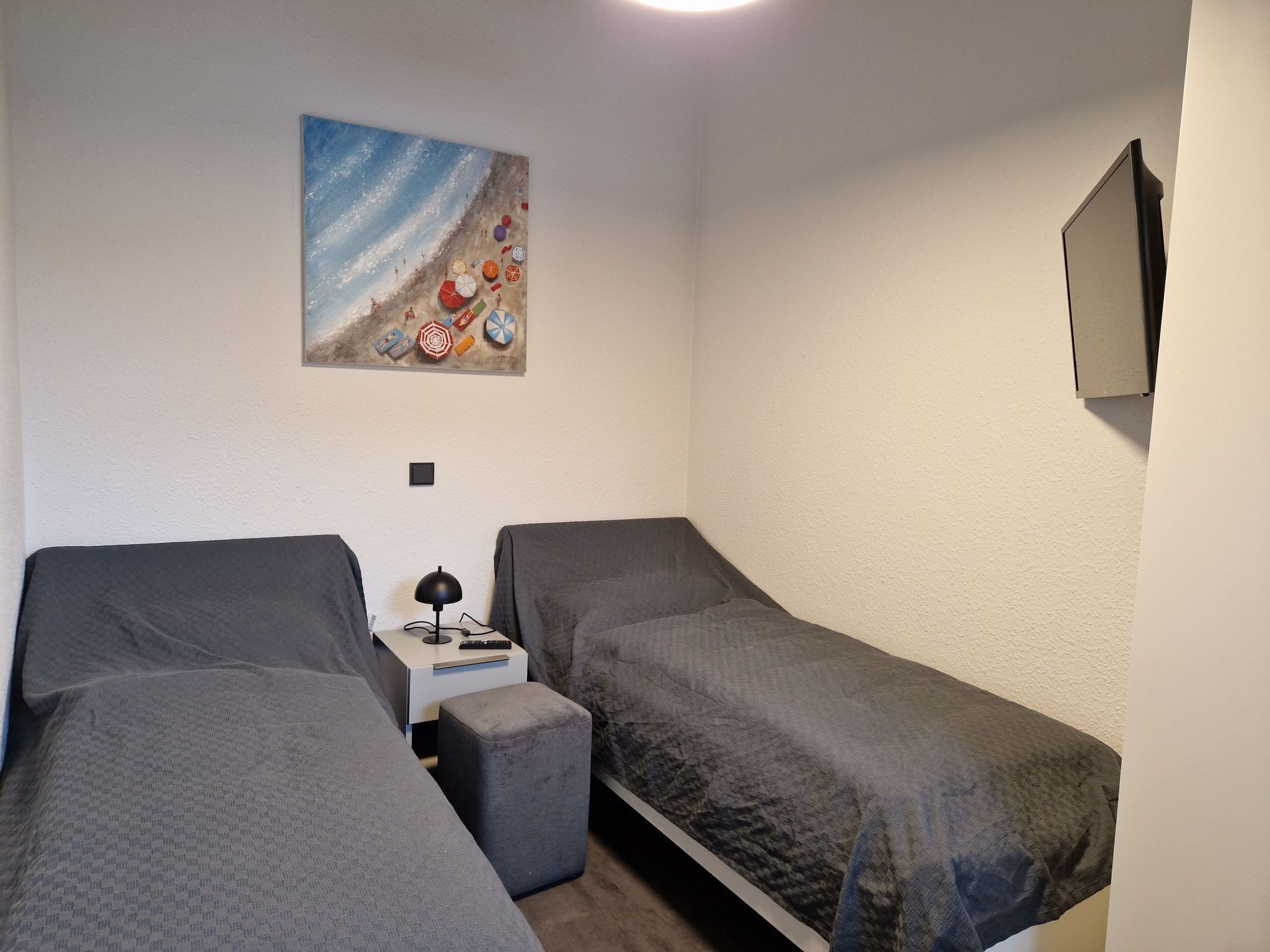 Une petite pièce, bien rangée, avec deux lits simples, une petite table avec une lampe et un tableau repr ésentant une scène de plage accroché au mur.