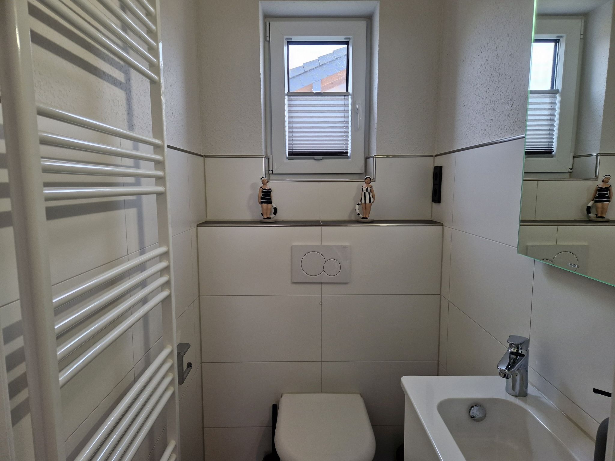 Un baño pequeño y moderno con inodoro, lavabo y ventana sobre el inodoro. Hay dos figuritas decorativas sobre el mostrador del lavabo.
