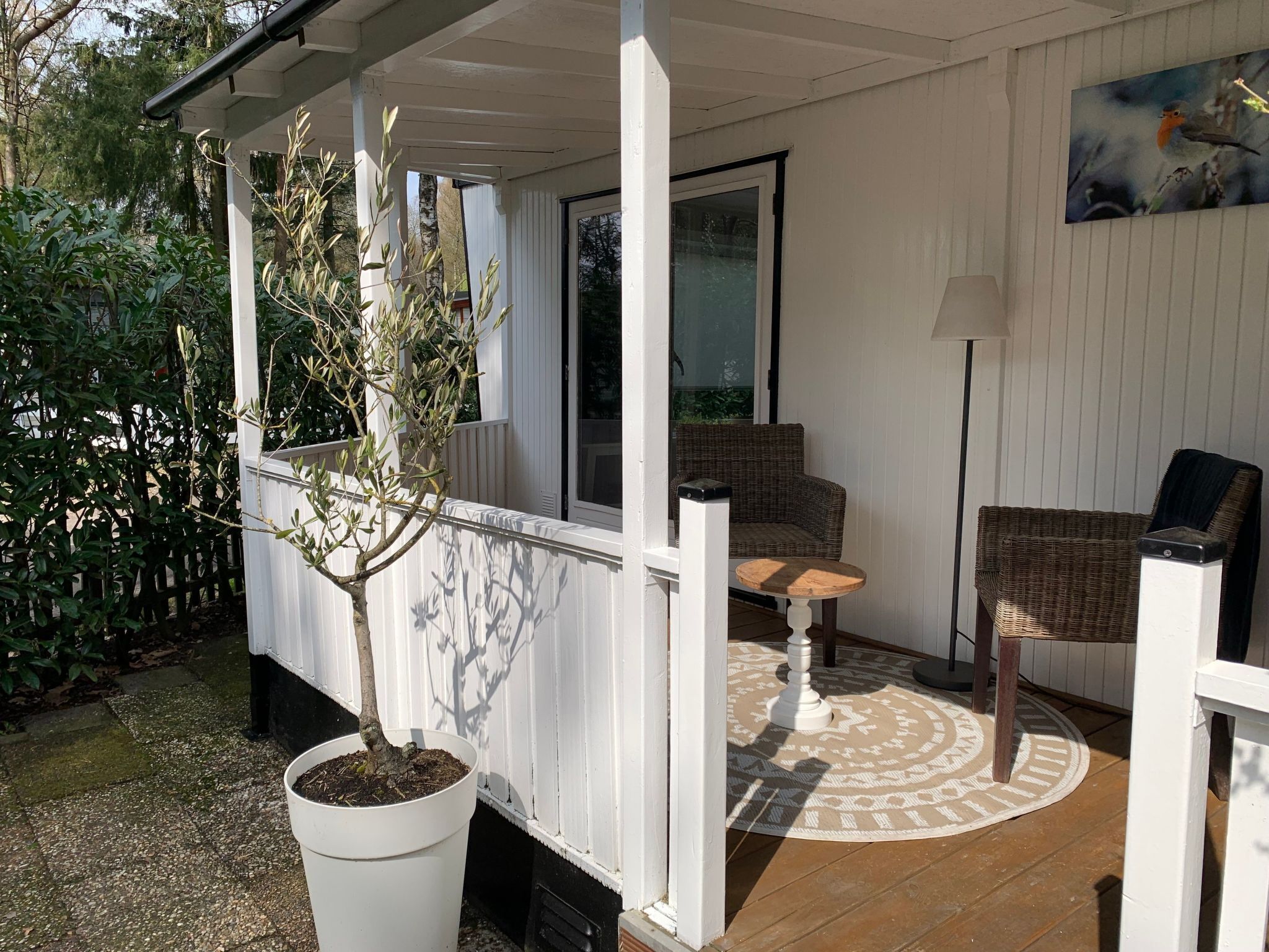 A covered patio area with a white railing, a potted tree, a wicker chair, a small table, and a floor lamp.