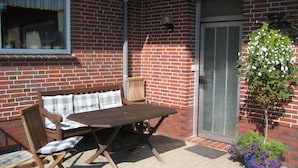 Eine Terrasse mit Holztisch und -stühlen, einer Ziegelwand, einer Schiebetür und Kübelpflanzen.