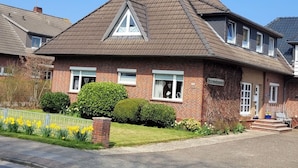 Ein zweistöckiges Haus mit Satteldach, einem sichtbaren Schornstein und einem kleinen Vorgarten mit gelben Blumen.