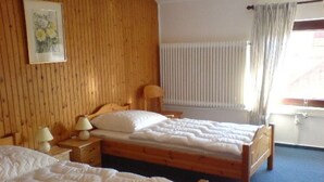 Ein Zimmer mit zwei Holzbetten, einem Holz-Kopfteil, einem Nachttisch mit Lampe, einem Fenster mit Vorhängen und einer Heizung.