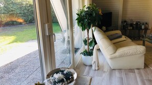Un salon moderne doté d’une grande porte-fenêtre coulissante donnant sur un jardin, d’un canapé beige, d’une plante en pot et d’une table basse en verre.