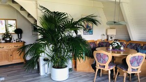 Un coin repas avec une table et des chaises en bois, une grande plante en pot et un escalier en arrière-plan.