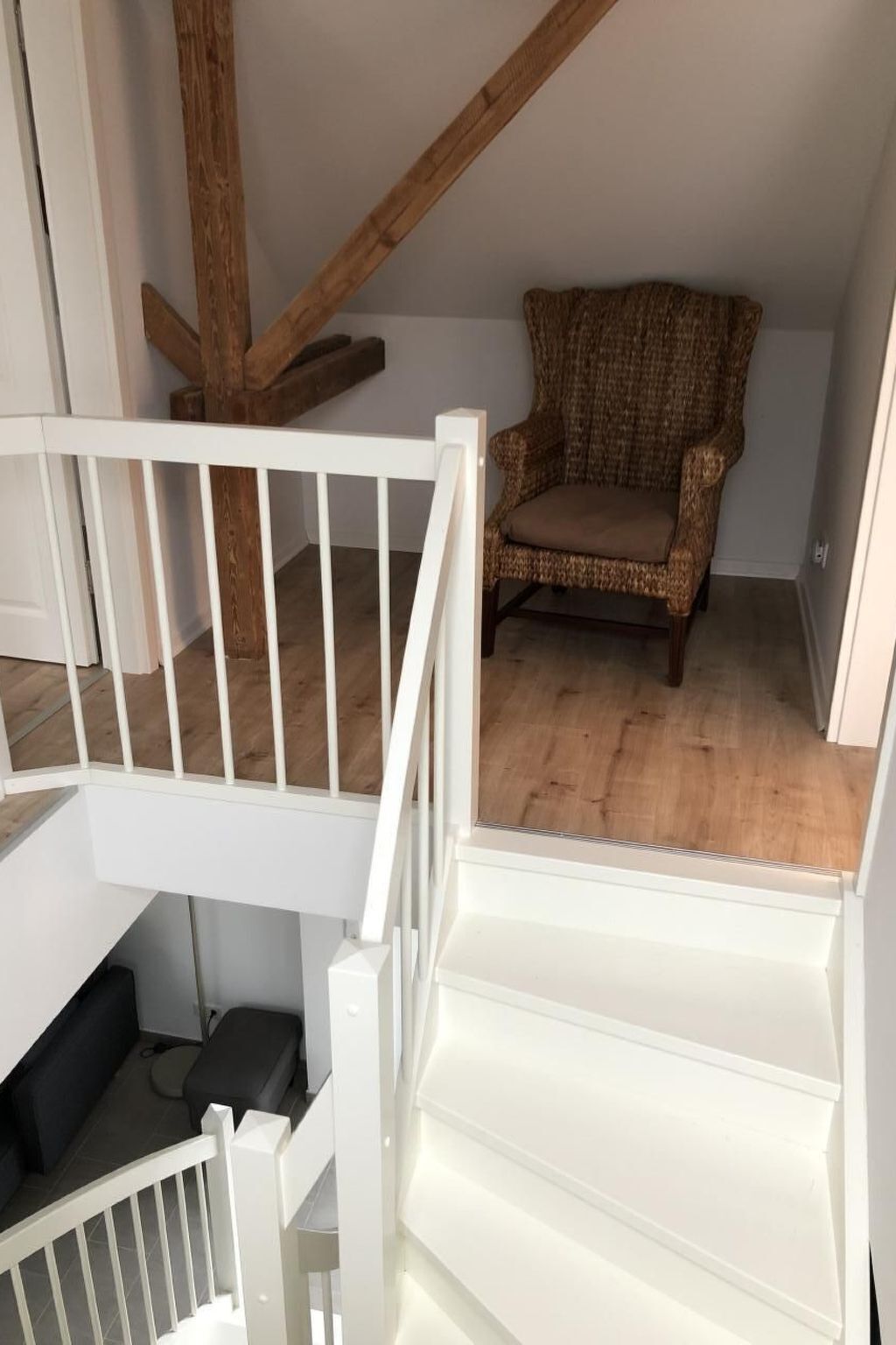 Une pièce avec un escalier blanc, une chaise en osier et une poutre en bois au plafond.