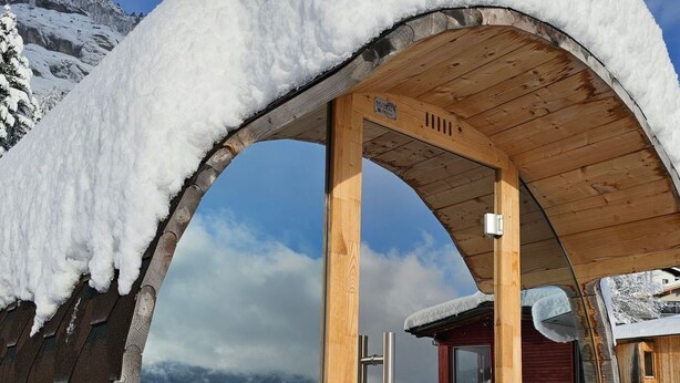 Ein mit Schnee bedecktes Holzgebäude mit rundem Fenster vor einer verschneiten Berglandschaft.