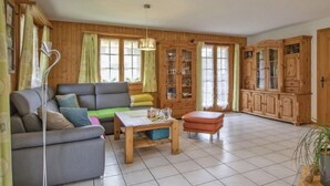 Een gezellige woonkamer met een grijze bank, een houten salontafel en een groene fauteuil. Er staan houten kasten en een groot raam met gordijnen.