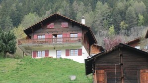 Un chalet en bois à deux étages, doté d’un balcon, situé au pied d’une colline boisée.
