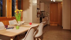 A dining area with a table, chairs, and a vase of flowers. A kitchen is visible in the background with cabinets and a stove. - Chalet 9 by Interhome (Innertkirchen)