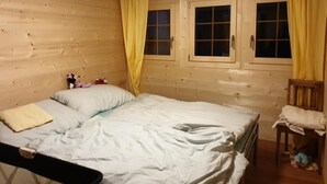 Ein Zimmer in einer Holzhütte mit einem Bett, zwei mit Vorhängen versehenen Fenstern, einem kleinen Stuhl und einem Heizkörper.