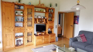 Ein Wohnzimmer mit Glastisch, grauem Sofa, Holzregalen mit Büchern und Dekoration, Fernseher und einer Pendelleuchte.
