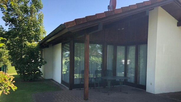 A covered patio area with a table and chairs, surrounded by glass walls and a brick floor. - K5 Haus in Eckwarderhörne mit Terrasse und Garte by Interhome (Burhave)