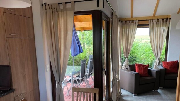 A room with a wooden dining table, chairs, a sofa with red cushions, and a TV on a cabinet. There is a sliding glass door leading to an outdoor area with a table and chairs. - Vacation home  by Interhome (Burhave)