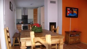 Un coin repas avec une table et des chaises en bois, une cuisine à l’arrière-plan et un téléviseur accroché au mur.