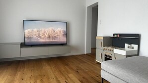 A modern living room with a flat-screen TV, wooden flooring, a sofa, and a kitchenette. - Sunnuhof Lodge by Interhome (Grächen)
