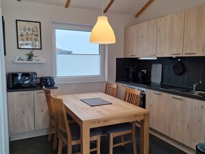 A compact kitchen with wooden cabinets, a black countertop, a dining table with chairs, a hanging light fixture, and a window with blinds. - G4 Nordsee-Ferienhaus mit Sauna und Garten by Interhome (Burhave)