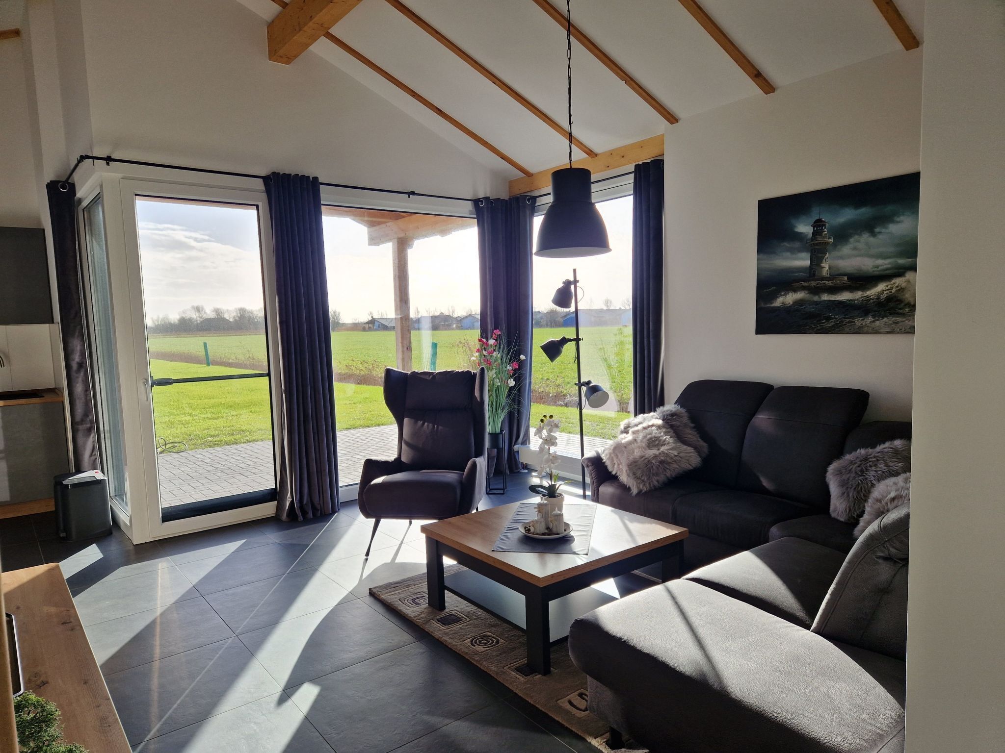 Ein modernes Wohnzimmer mit einer Couch, einem Sessel und einem Couchtisch. Es gibt ein großes Fenster mit Vorhängen, eine Stehlampe und ein Bild an der Wand.