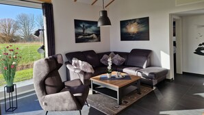 Ein stilvolles Wohnzimmer mit Sofa, Sesseln und Couchtisch. An den Wänden hängen zwei Bilder, eine Zimmerpflanze und ein Blick ins Freie durch ein großes Fenster.
